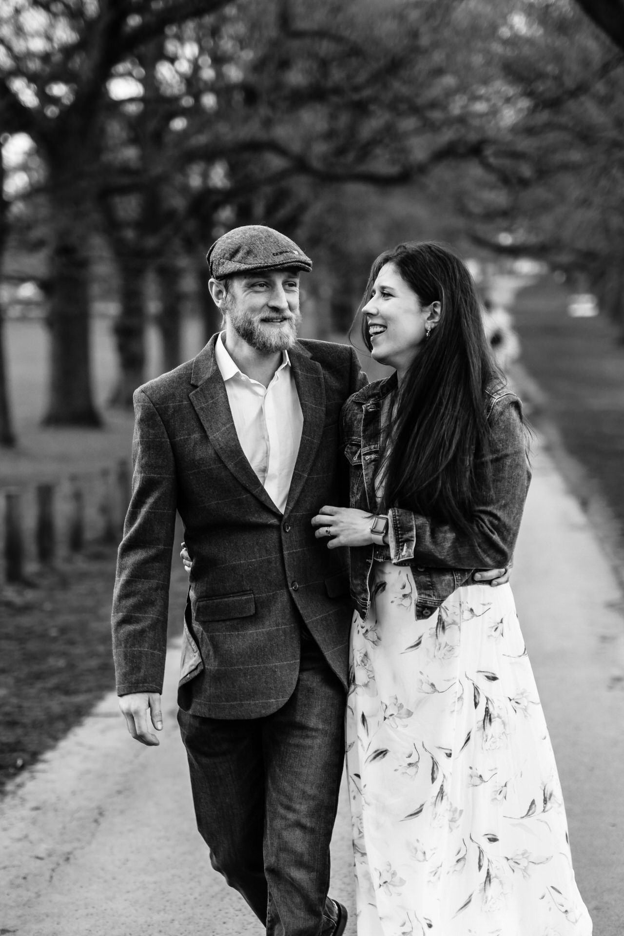 A black and white portrait of a couple sharing a joyful moment on a tree-lined path.