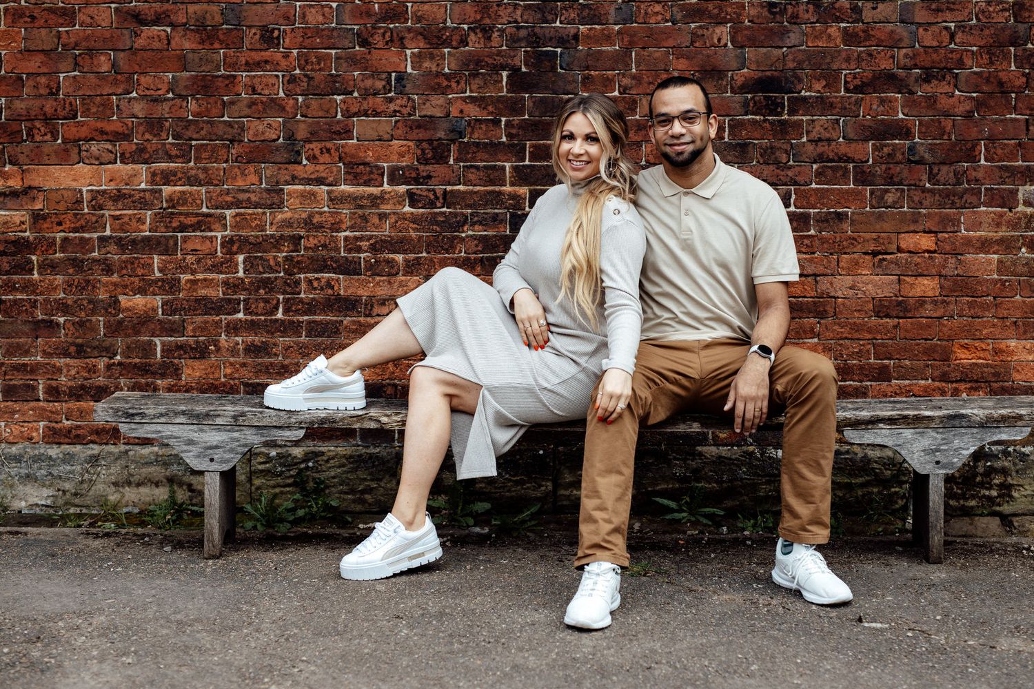 A couple in coordinated outfits poses casually against a weathered brick wall.