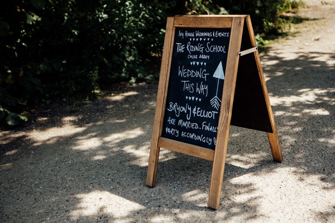Wooden A-frame chalkboard signs in dappled sunlight at an outdoor wedding venue.