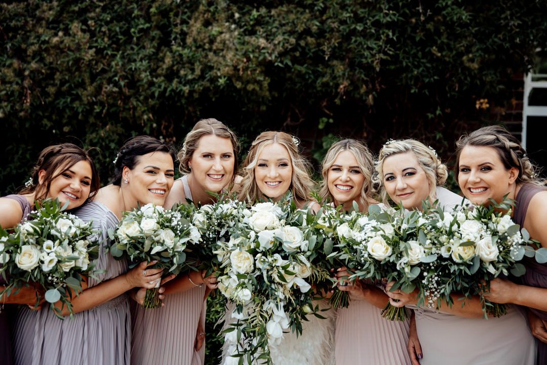 Wedding party holding white and green floral bouquets against rustic backdrop.