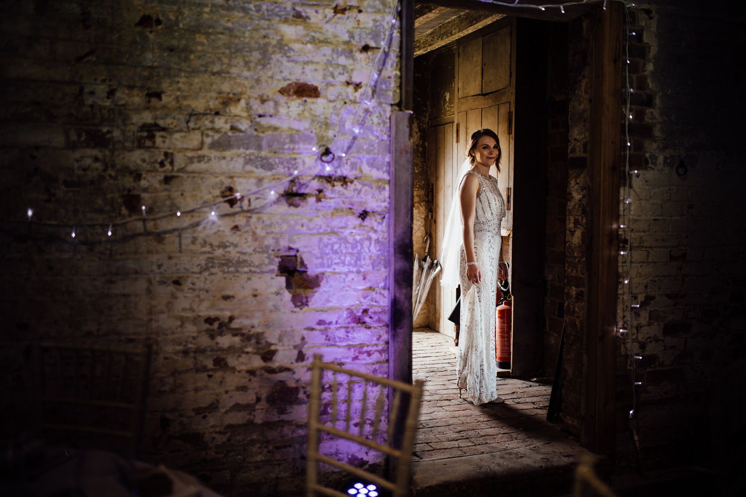 A figure in white stands in a dramatic doorway with purple lighting in an old brick building.