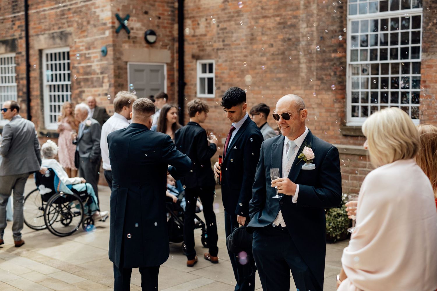 Wedding guests mingling outside a historic brick building with large windows.