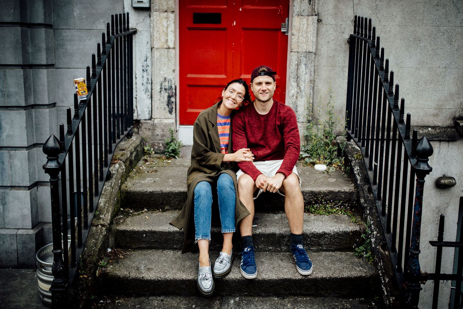 Couple sits on concrete steps in front of red door with black metal railings.