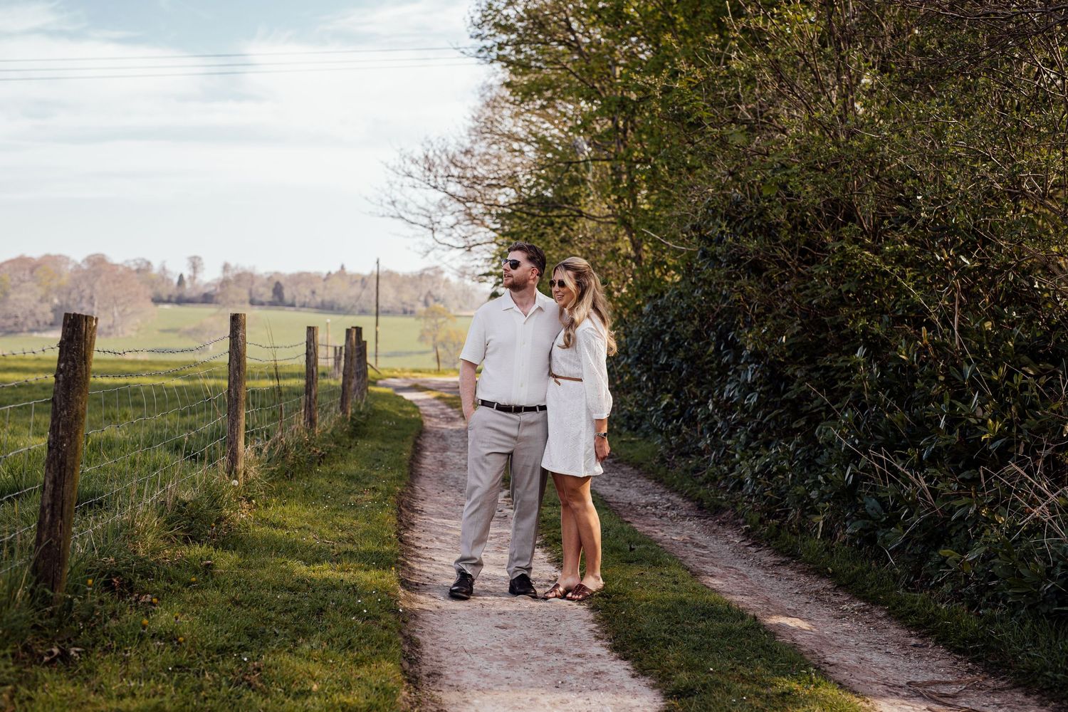 A couple strolls hand in hand along a tree-lined dirt path in a pastoral landscape.