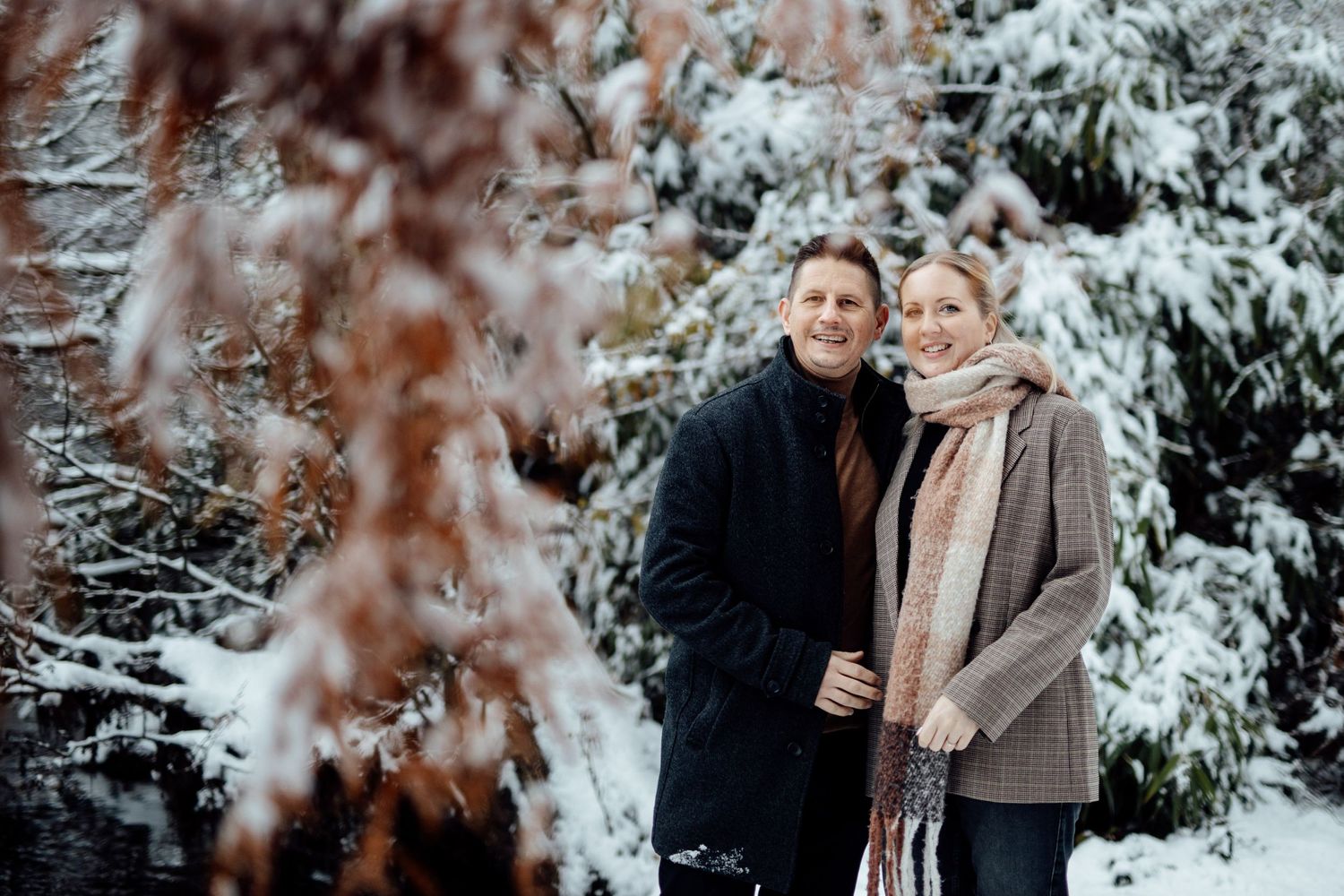 A couple stands close together in a snowy winter scene with snow-covered evergreen trees in the background.