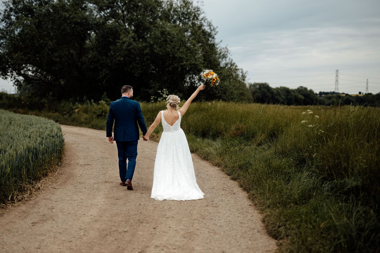 A couple walking down a dirt path through a countryside field on their wedding day.