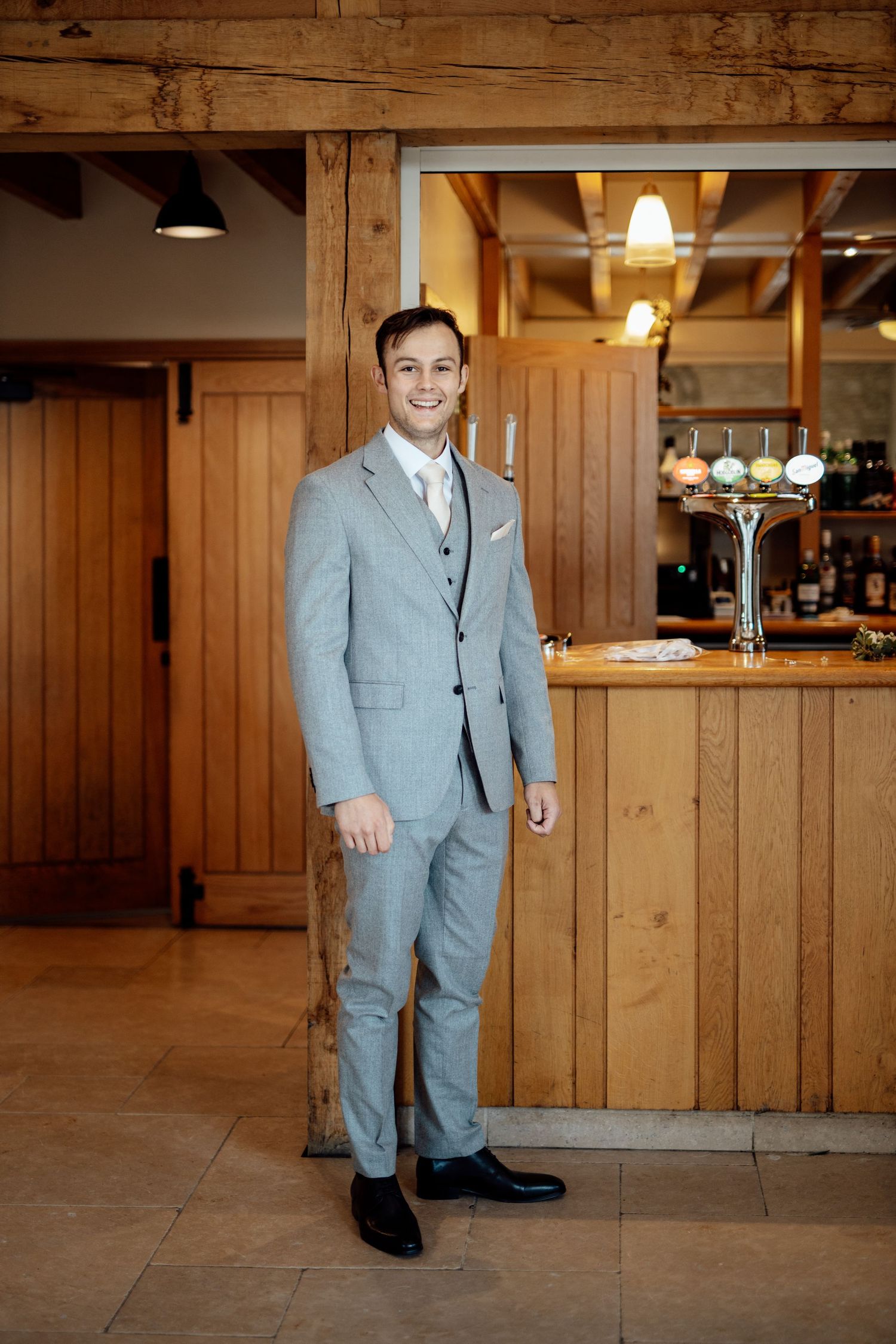 A young ring bearer in a light gray suit poses in front of a rustic wooden wall at a wedding venue.