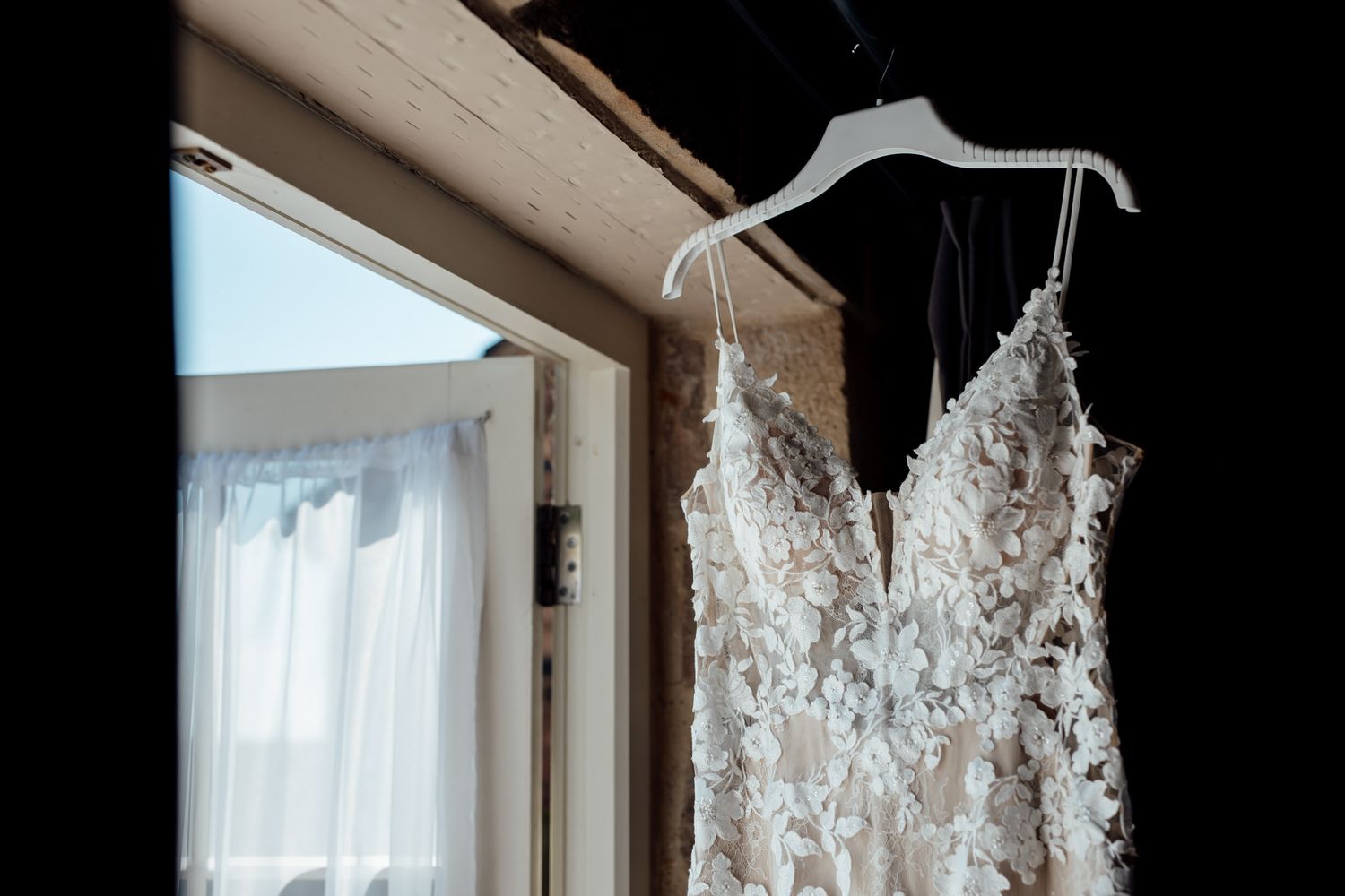 Elegant white lace wedding dress hanging in window light against dark background.