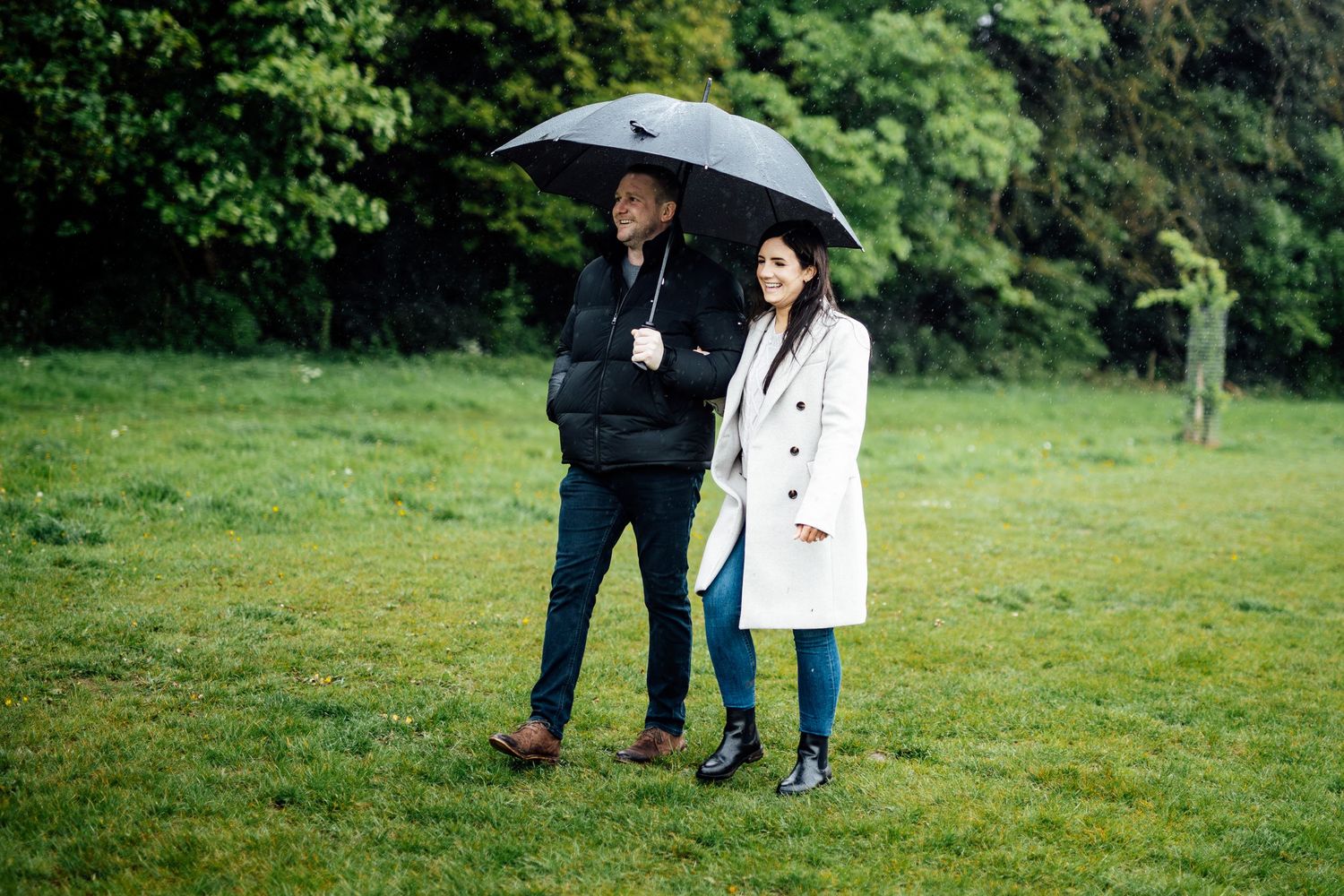 A couple walks together on a green path while one holds an umbrella on an overcast day.