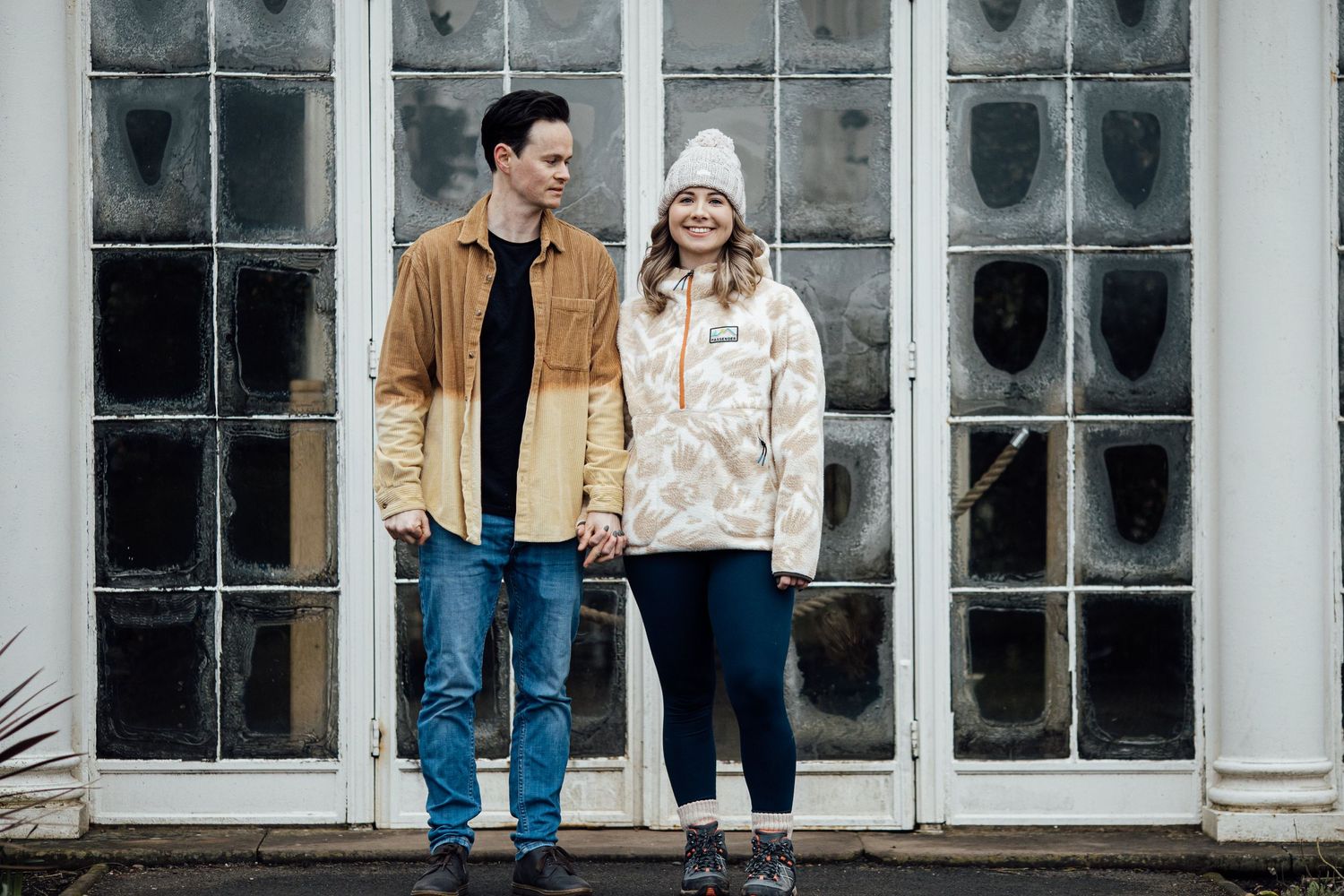 A couple standing together in casual outfits against a large window backdrop.