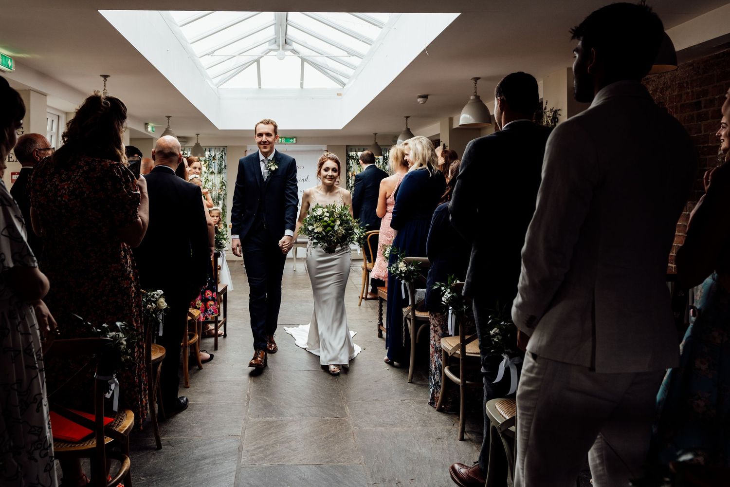 Wedding ceremony processional in venue with skylight and guests lining the aisle.
