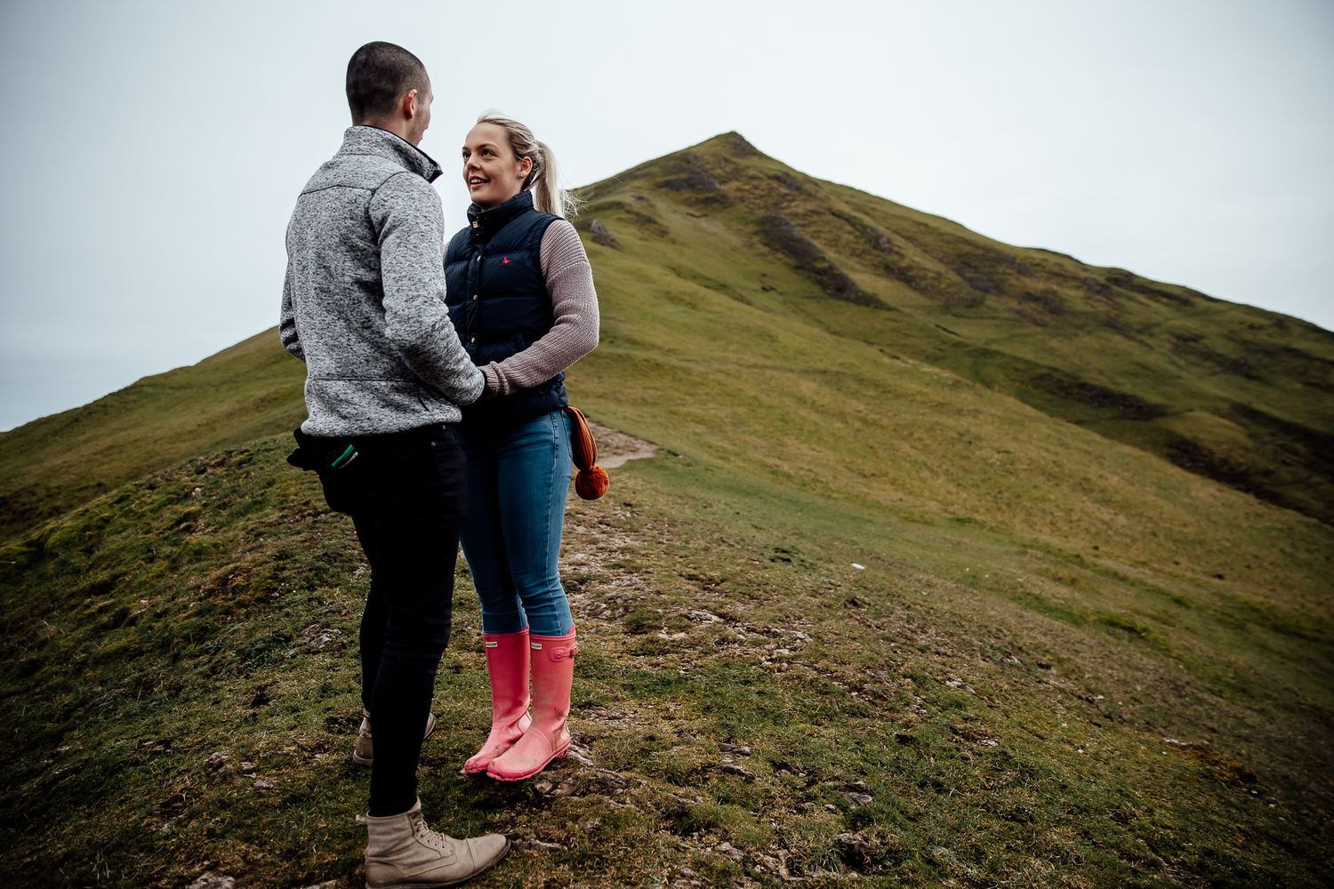 Two people walking together on a grassy hillside path against an overcast sky.