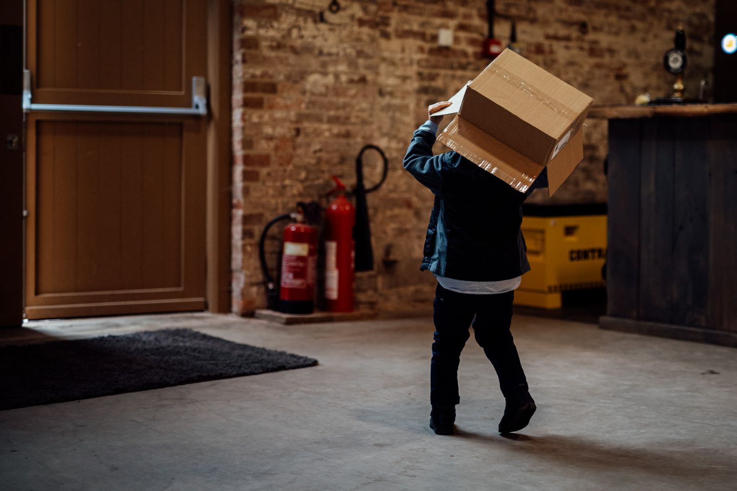 Person carrying a large cardboard box through an industrial space with brick walls and a fire extinguisher.