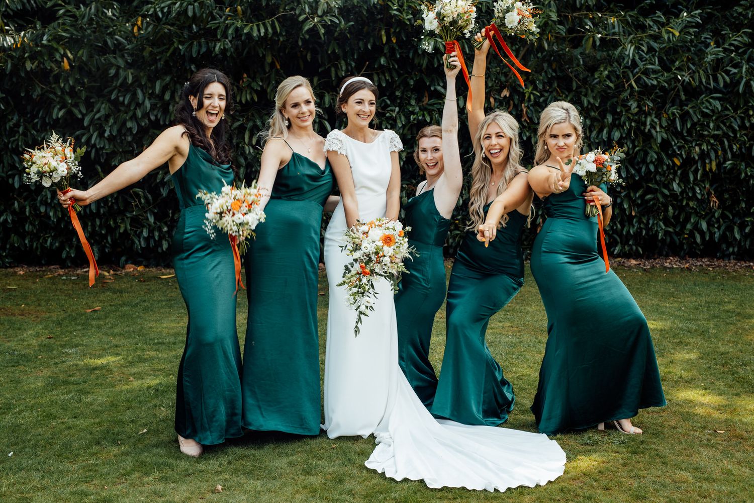 Bridal party posing together in emerald green dresses on a sunny lawn.