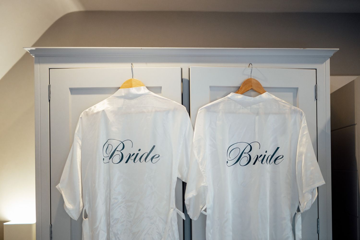 White bridal robes hanging on wooden hangers against neutral wall.