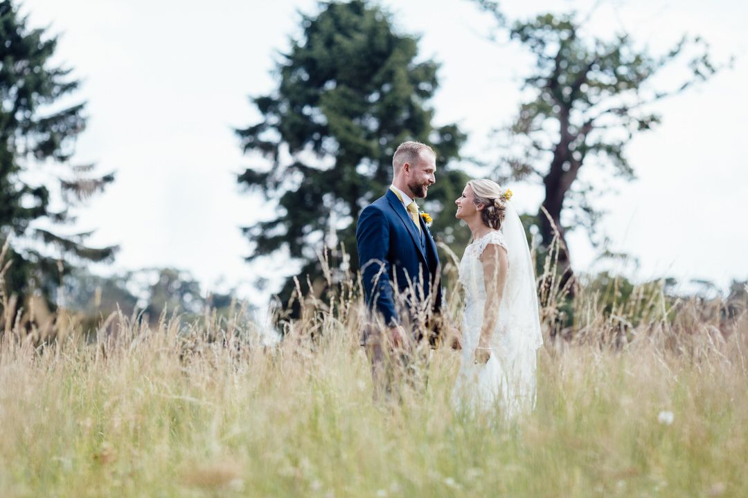 Wedding group standing in tall grass field with evergreen trees in background.