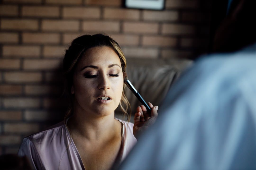 Professional makeup application session with soft lighting against a brick wall background.