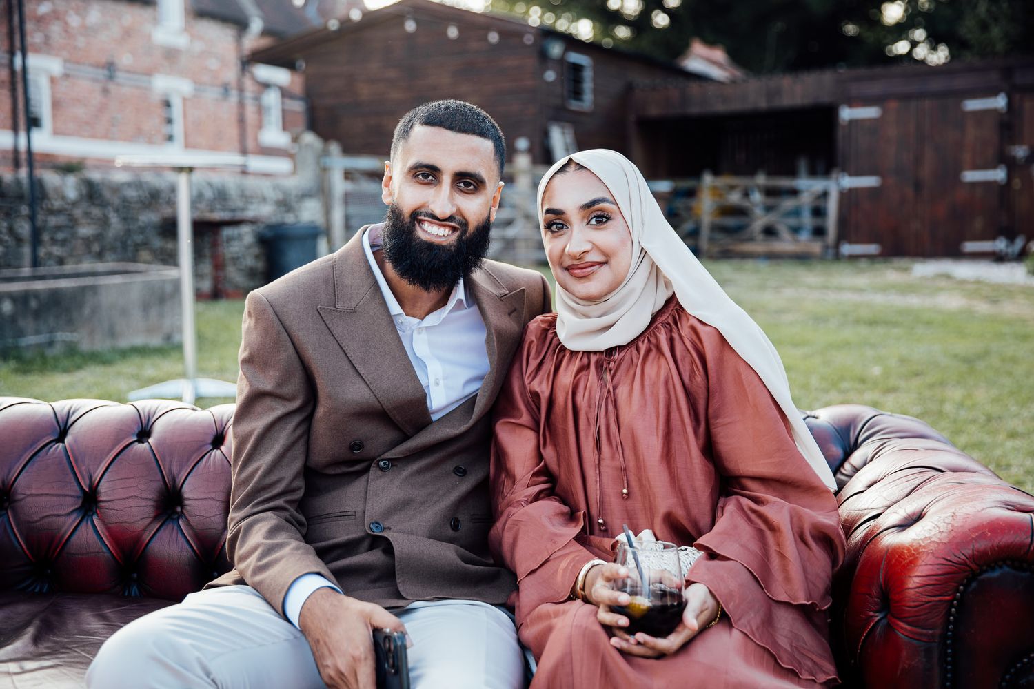 Couple in coordinated brown and rust colored outfits pose together on a vintage leather couch during engagement session.