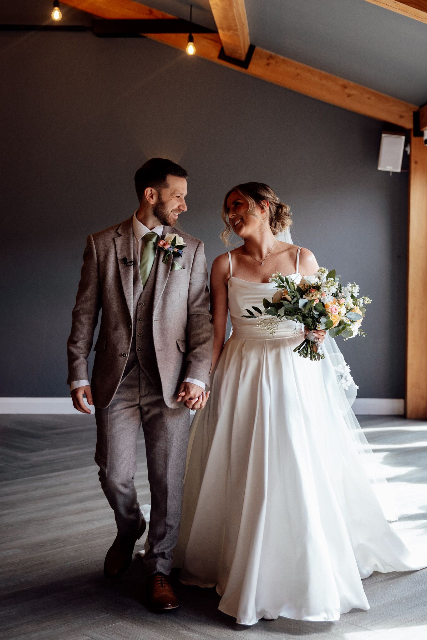 A couple in wedding attire walking together in an indoor venue with exposed wooden beams and dramatic lighting.