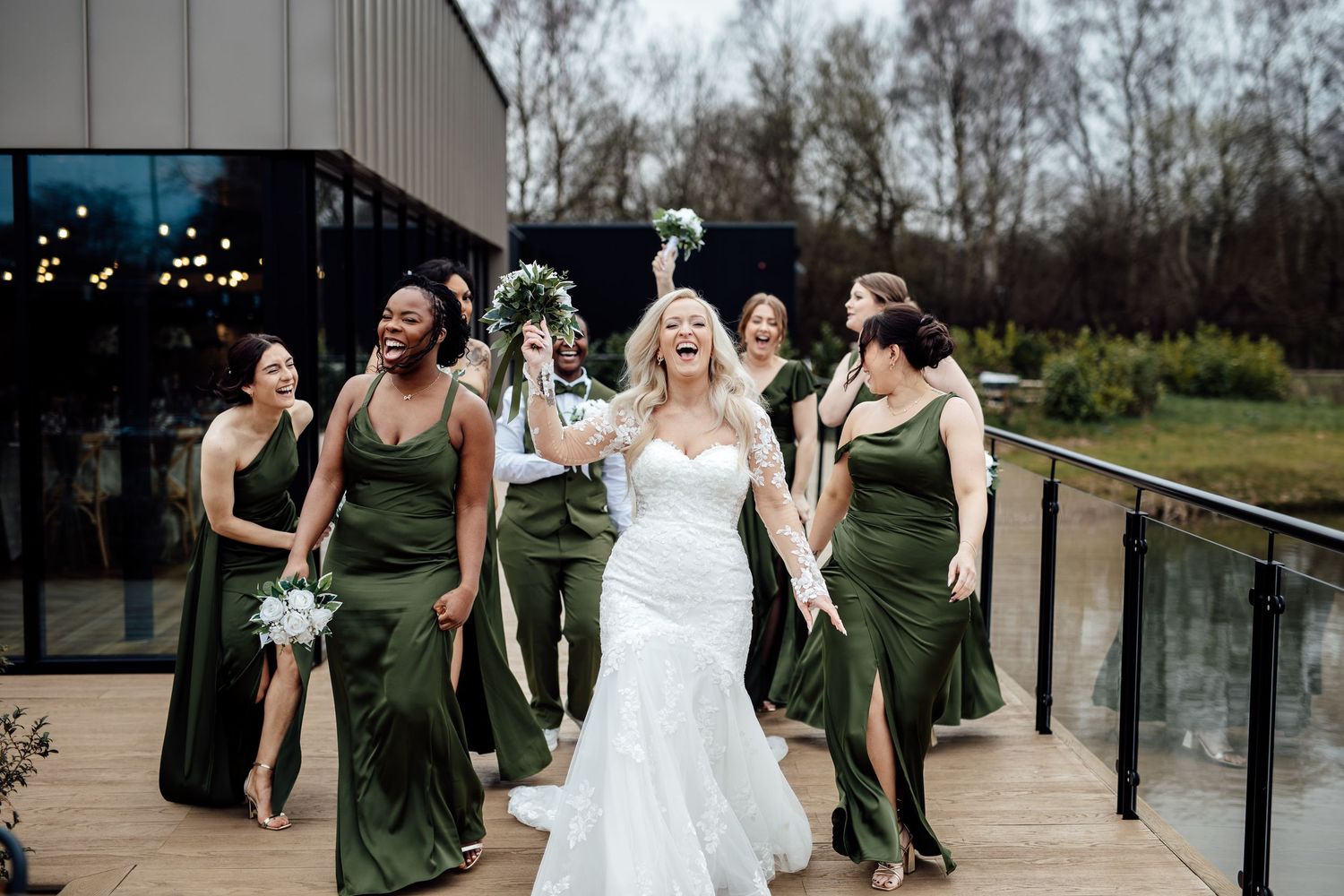 Bride in white gown celebrating with bridesmaids in olive green dresses outdoors.