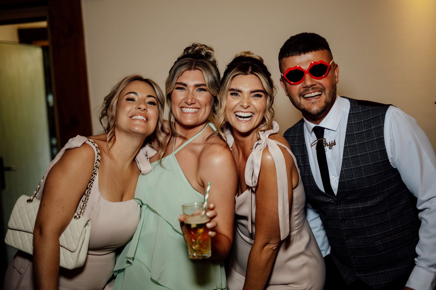 Four friends smile together at a wedding reception, with one man wearing fun red heart-shaped sunglasses.