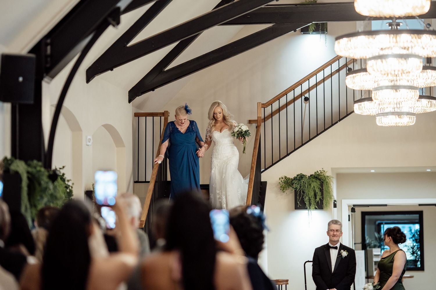 A bride descends a grand staircase with her mother as the groom watches during the wedding ceremony.