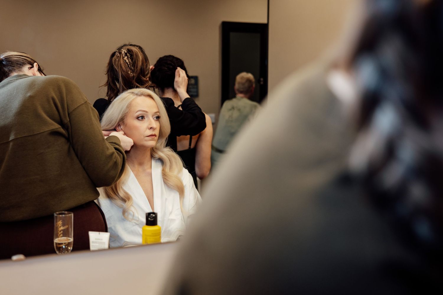 Blonde bride getting hair and makeup done by stylists in a bright wedding preparation room.
