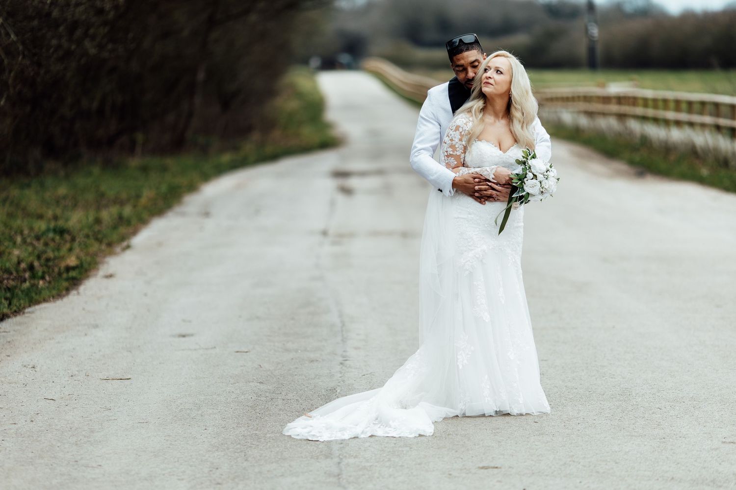 Bride and groom pose on a scenic road, bride in white lace gown holding bouquet, groom in white suit.