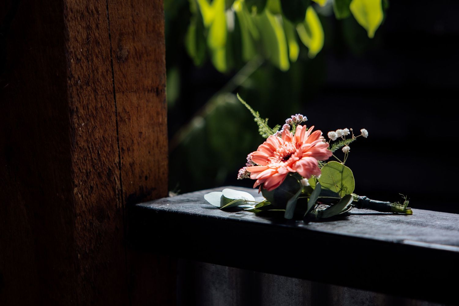 A delicate pink dahlia boutonniere resting on a dark wooden ledge with green foliage background.