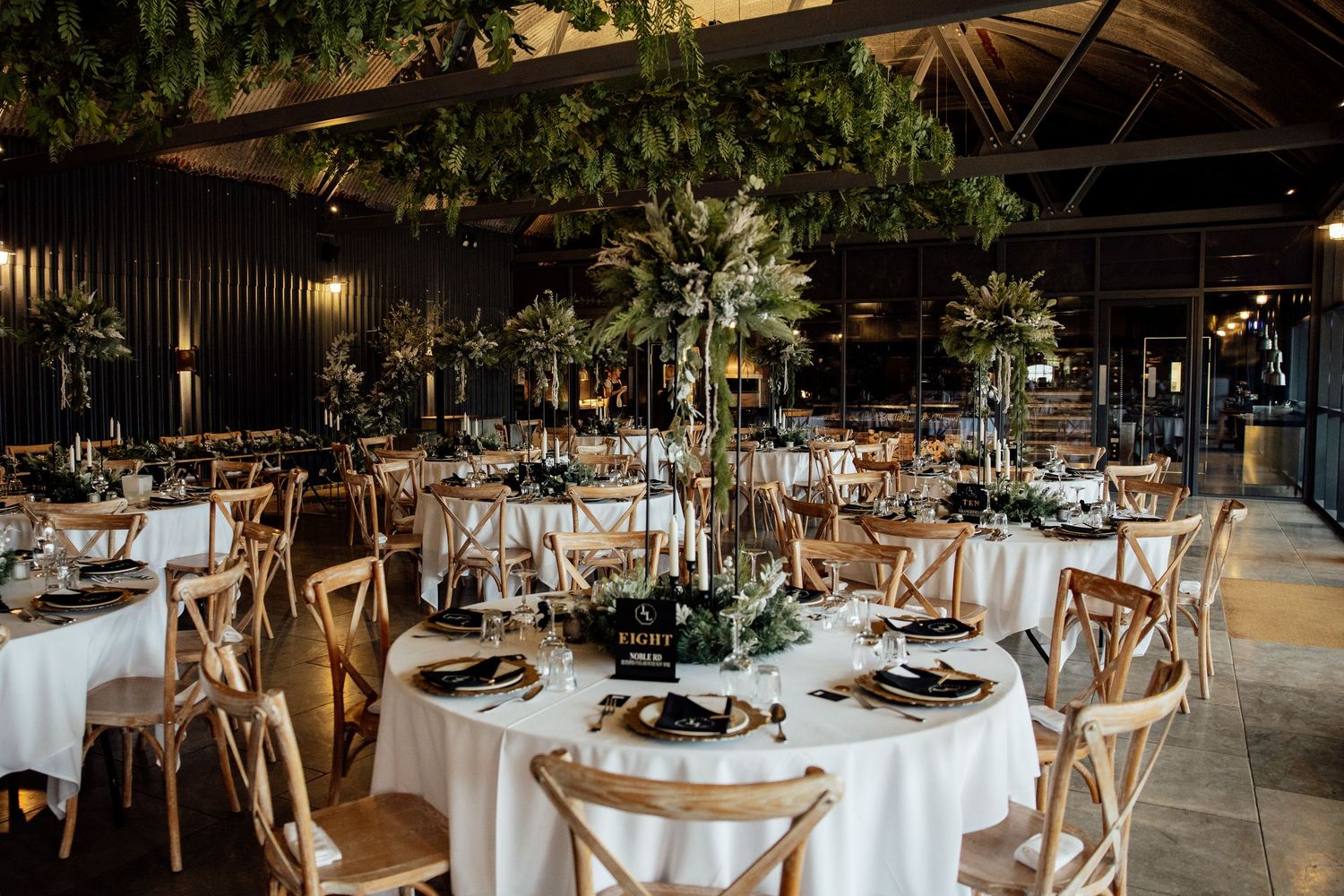 Elegant wedding reception tables decorated with greenery and gold accents in an industrial-style venue.
