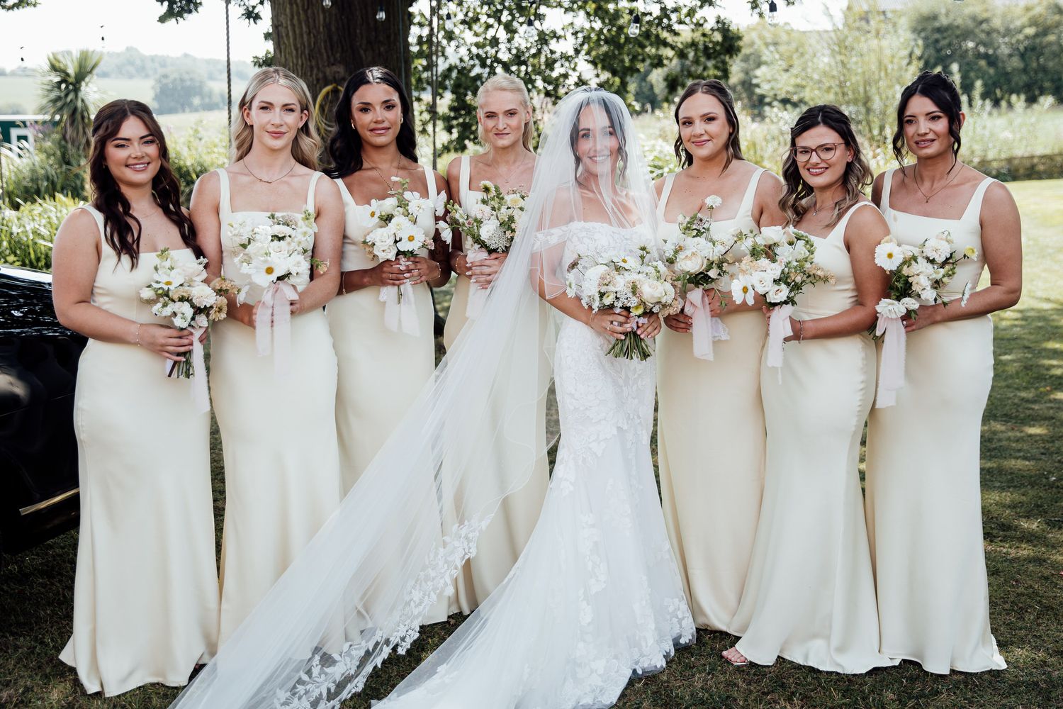 A bridal party in cream-colored dresses poses together outdoors holding bouquets.