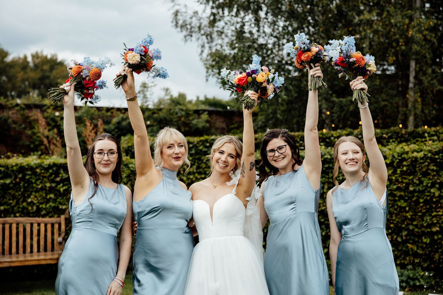 Bridesmaids in powder blue dresses hold colorful floral bouquets while posing outdoors.