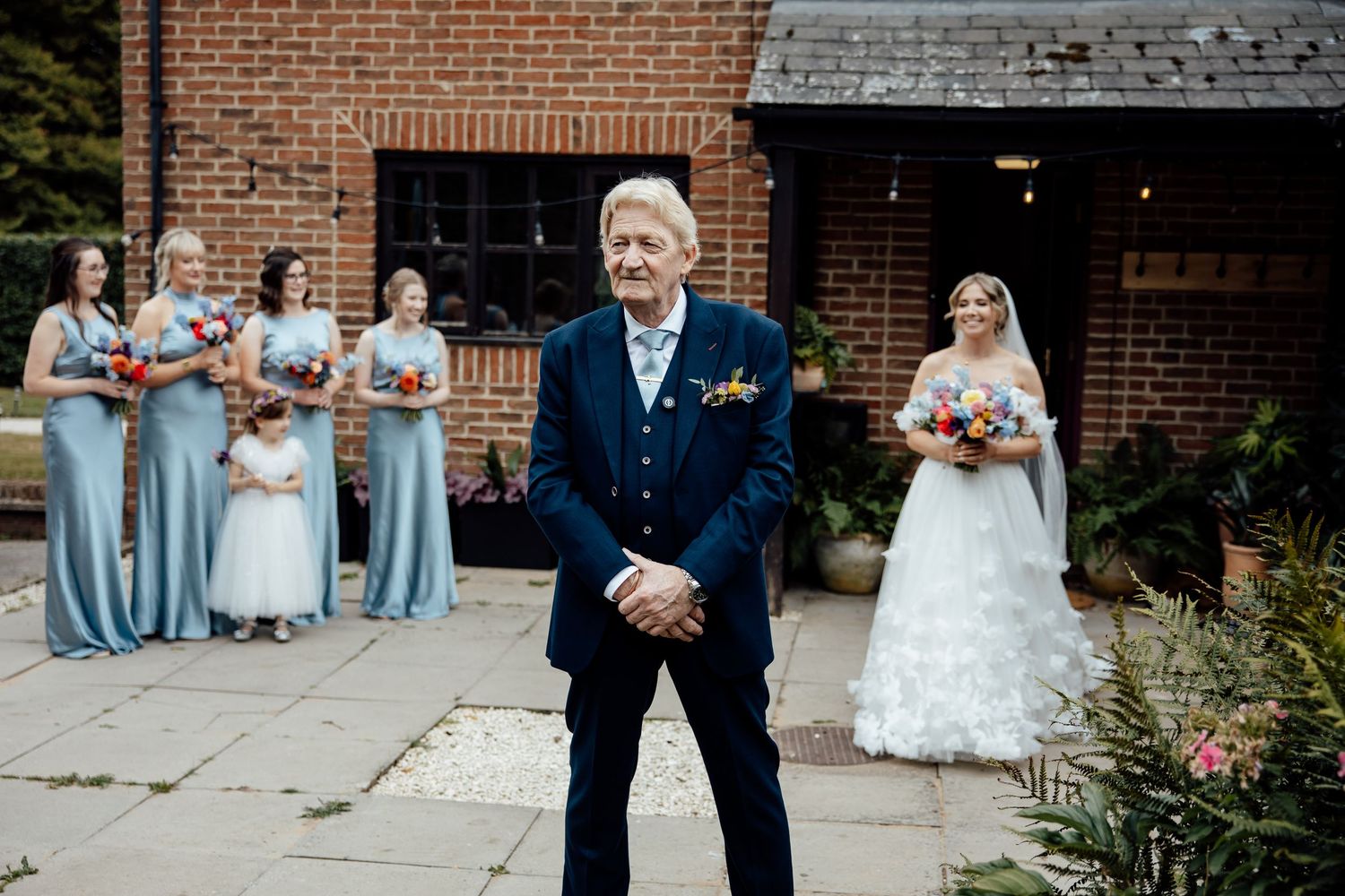 Wedding party in light blue bridesmaid dresses stands outside brick venue with navy-suited figure in foreground.