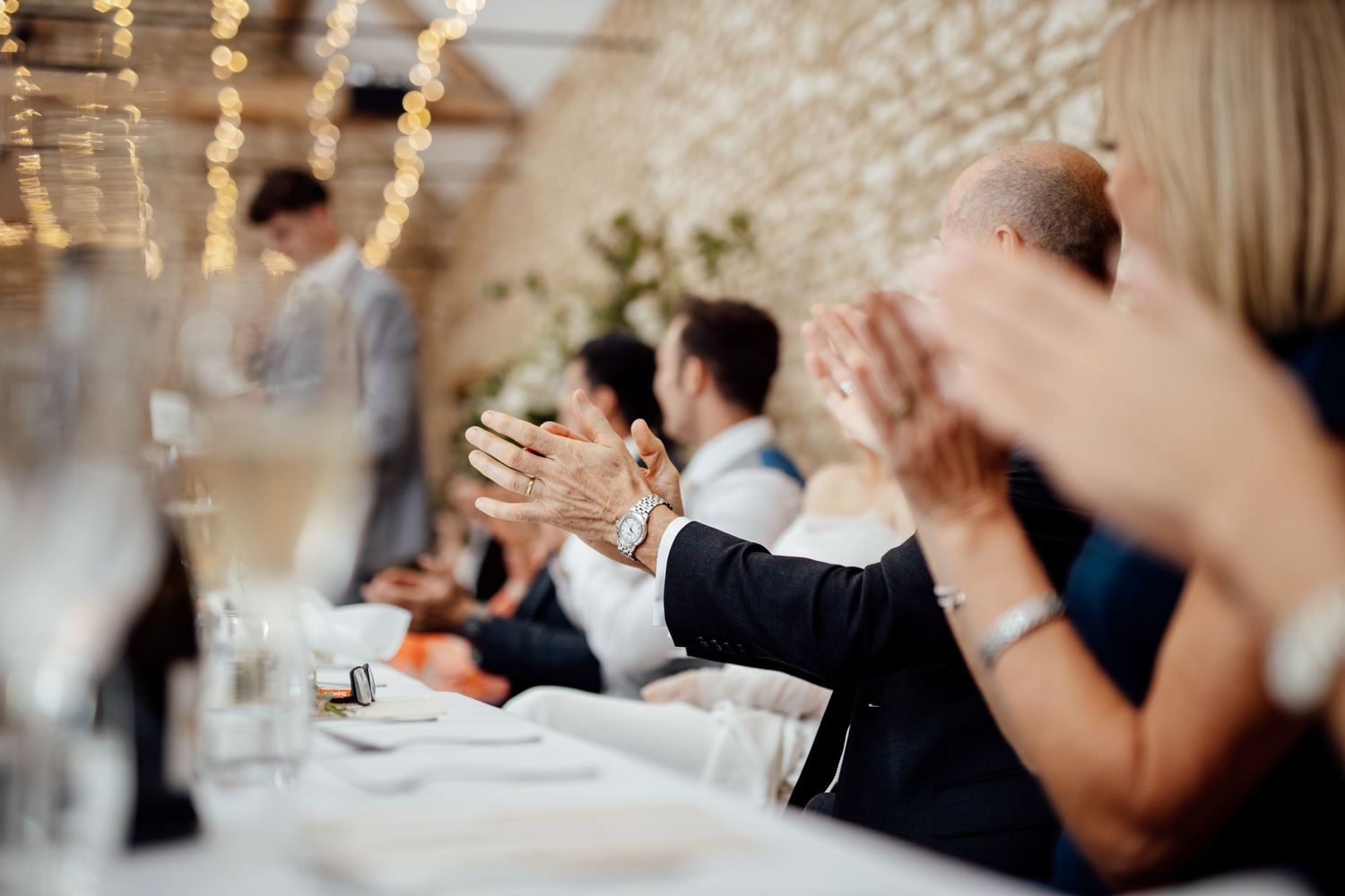 Wedding guests applauding during reception dinner at a stone-walled venue.