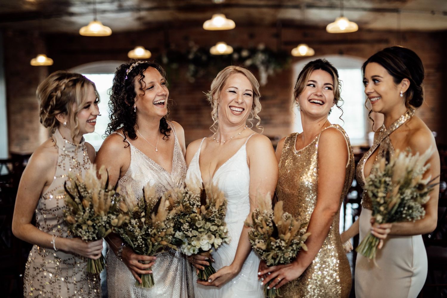 Bridesmaids in sparkly dresses hold bouquets while standing in rustic indoor venue.