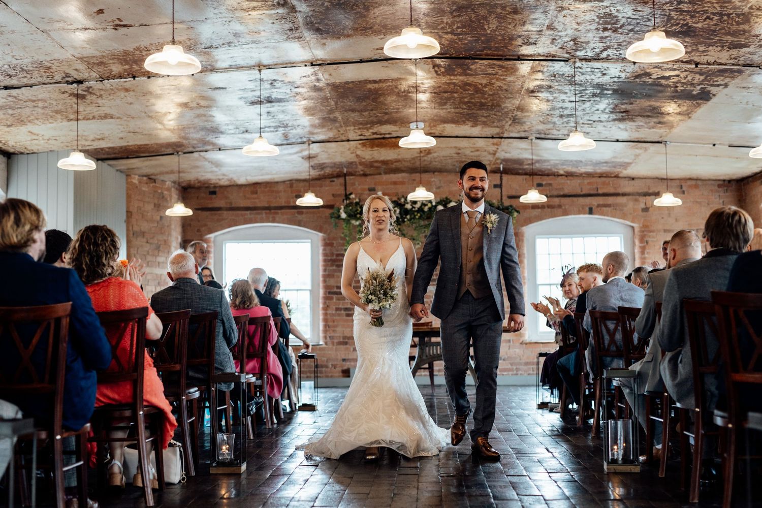 Couple walking down aisle in rustic brick venue with pendant lighting and seated guests.