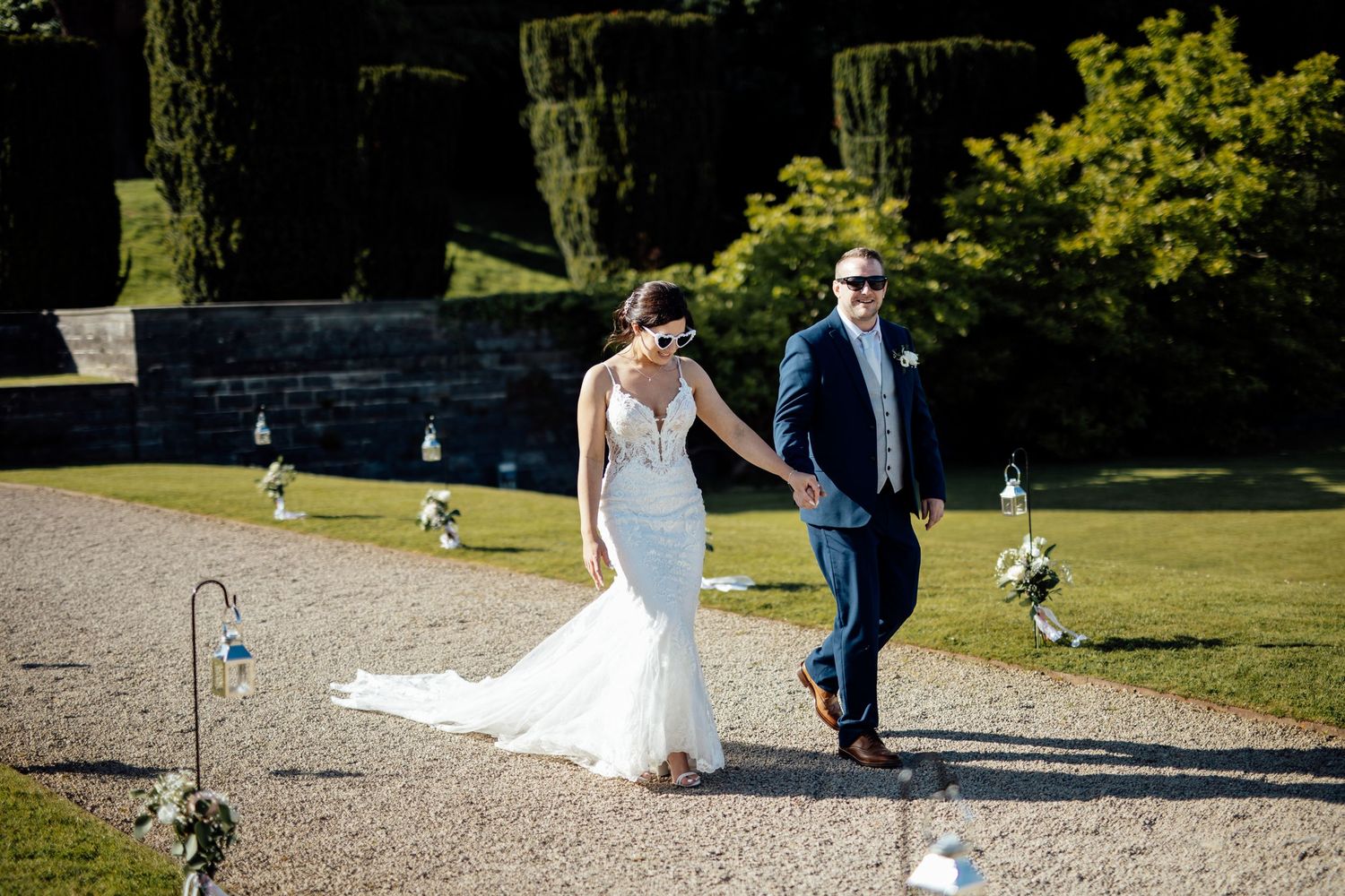 A couple walks along a garden path decorated with shepherd's hooks and floral arrangements on their wedding day.