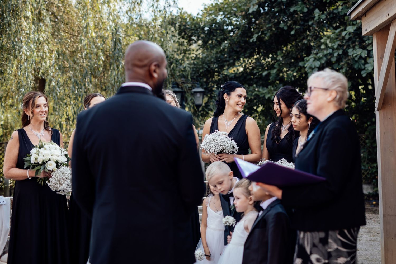 Wedding guests gather outdoors under trees for a ceremony at a scenic venue.