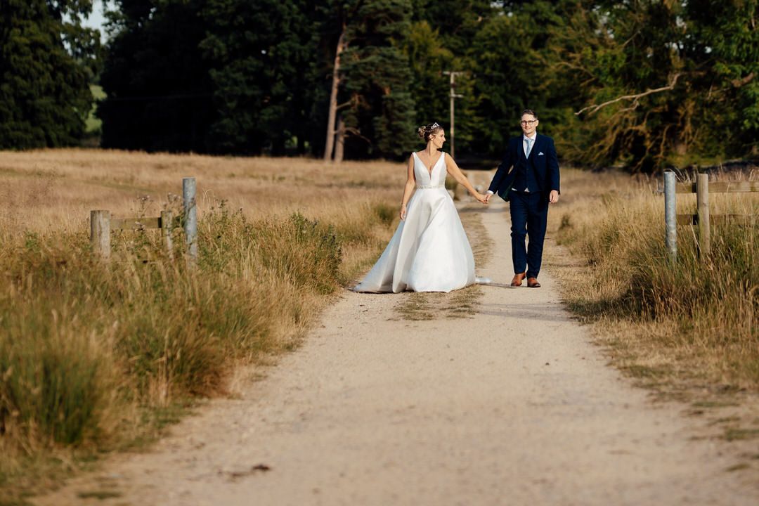 Couple walks down scenic dirt path surrounded by tall grass and trees.