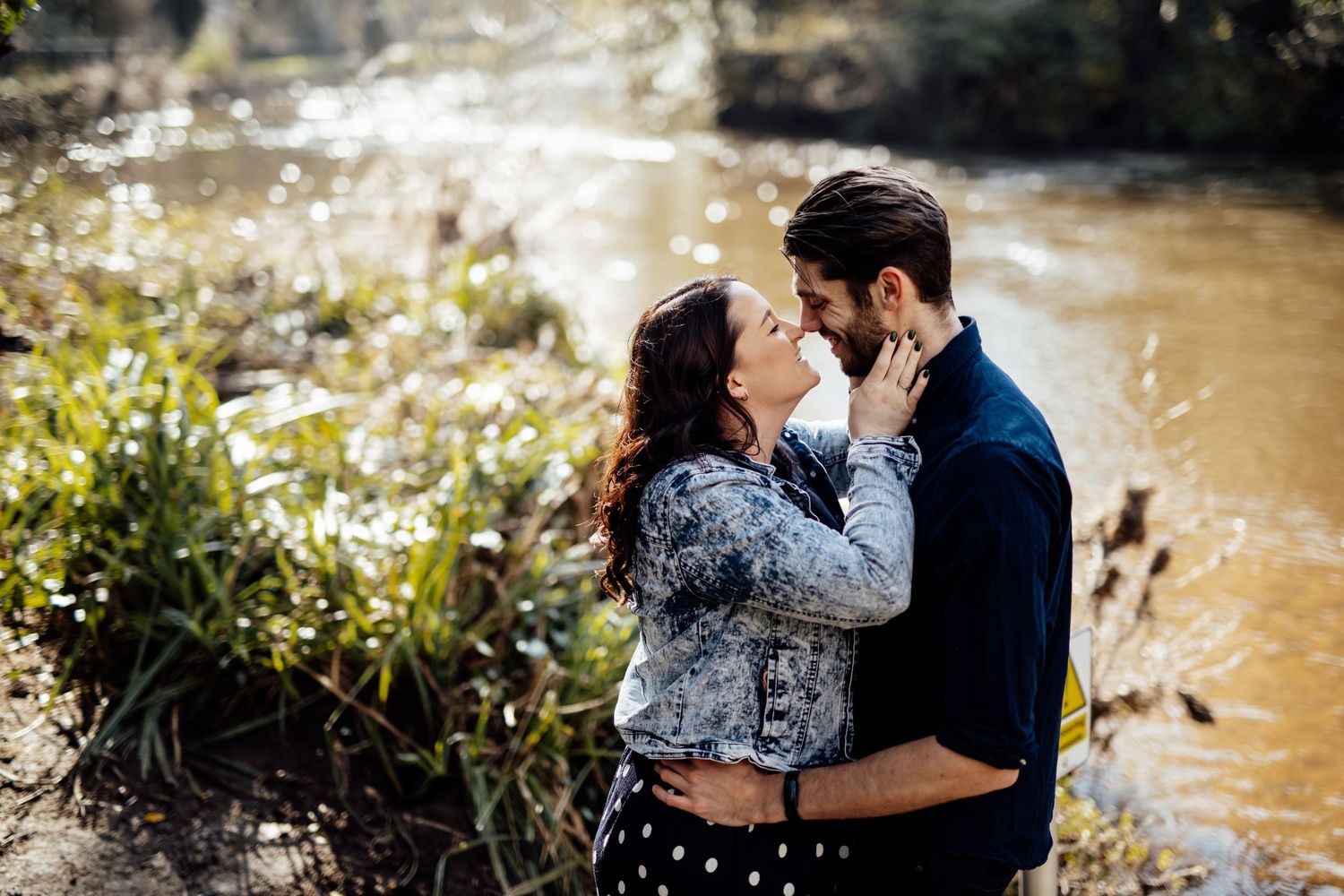 A couple shares an intimate embrace by a river with sunlight filtering through trees.