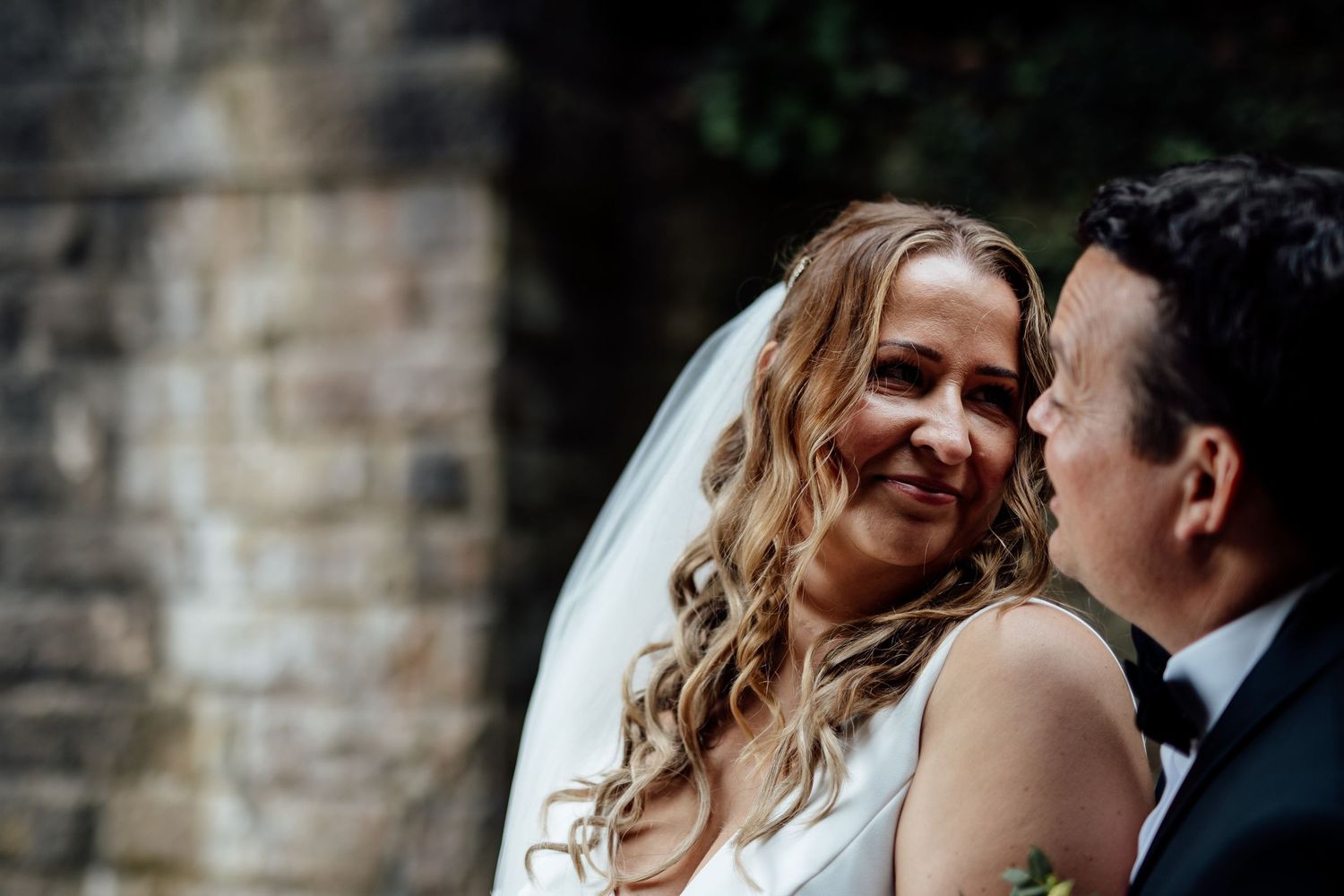 Romantic wedding couple embracing against stone wall backdrop.