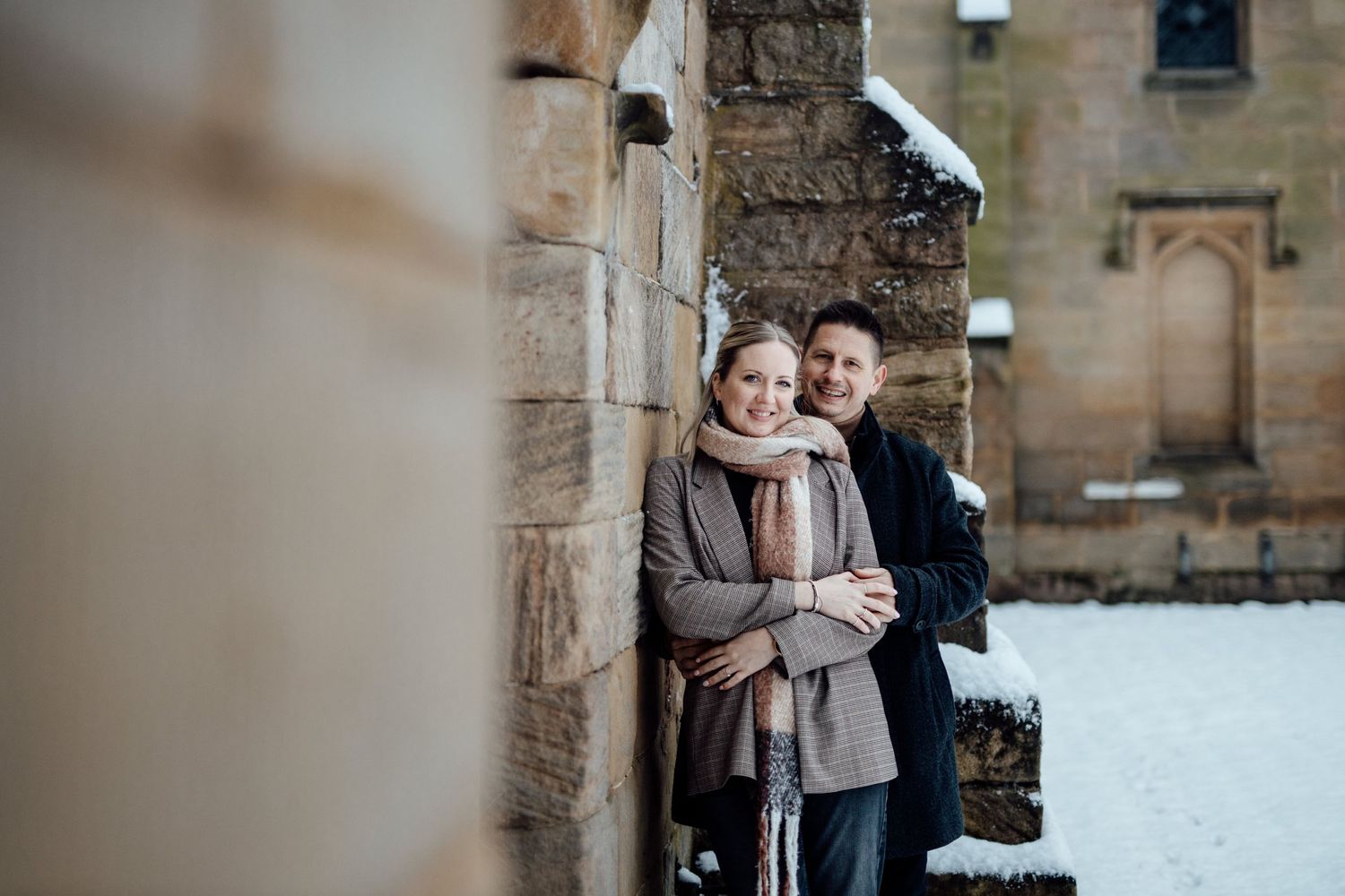 Couple standing close together against stone architecture in winter snow.