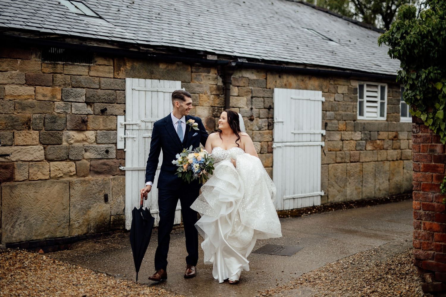 Couple walking along stone wall pathway with white door in rustic wedding setting.