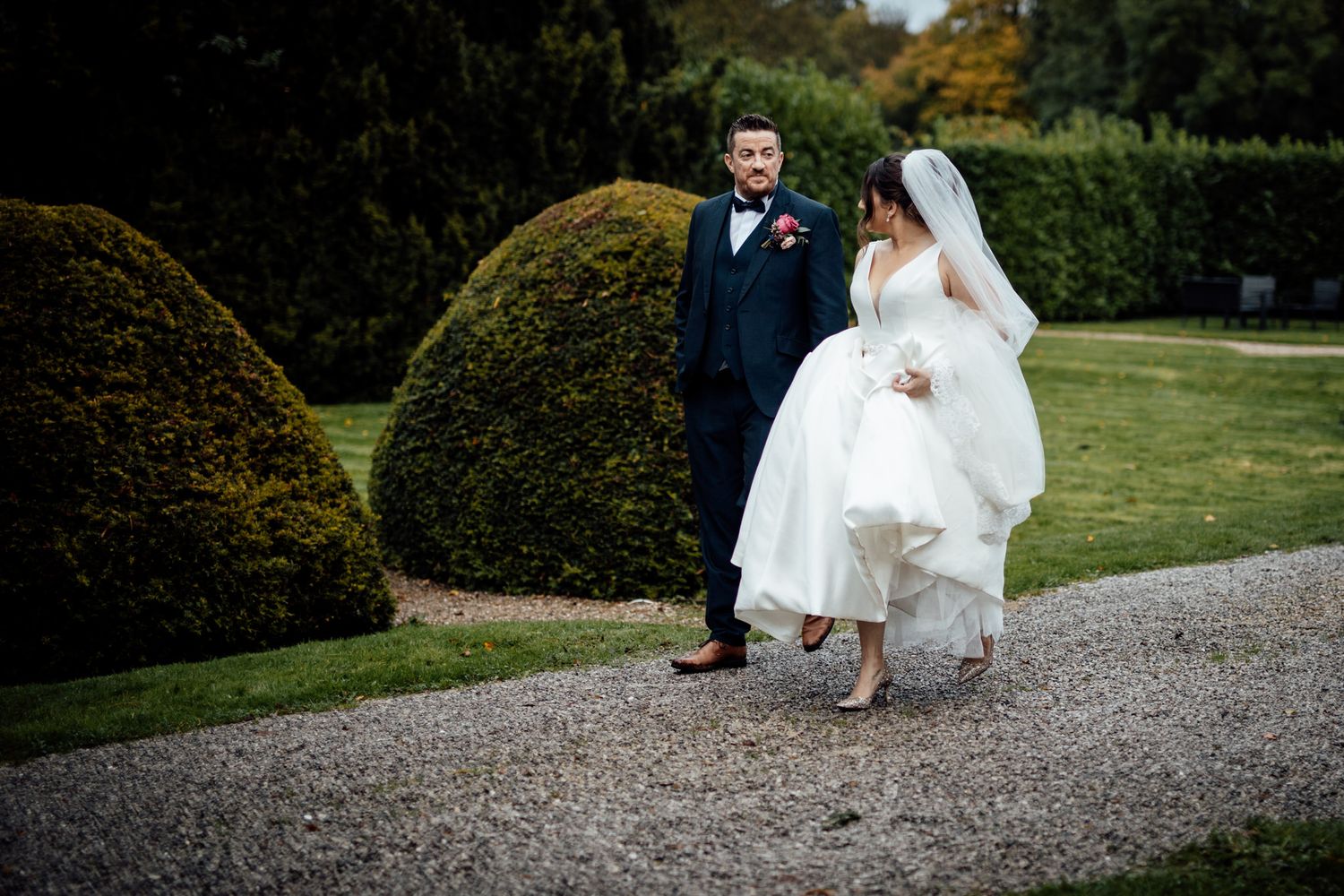 Wedding couple walking along a gravel path surrounded by manicured garden hedges.