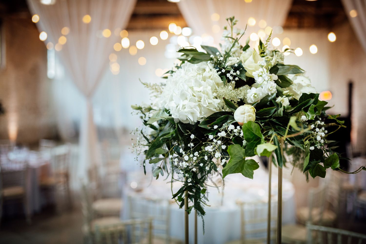 Elegant white floral centerpiece with greenery and string lights in wedding reception venue.