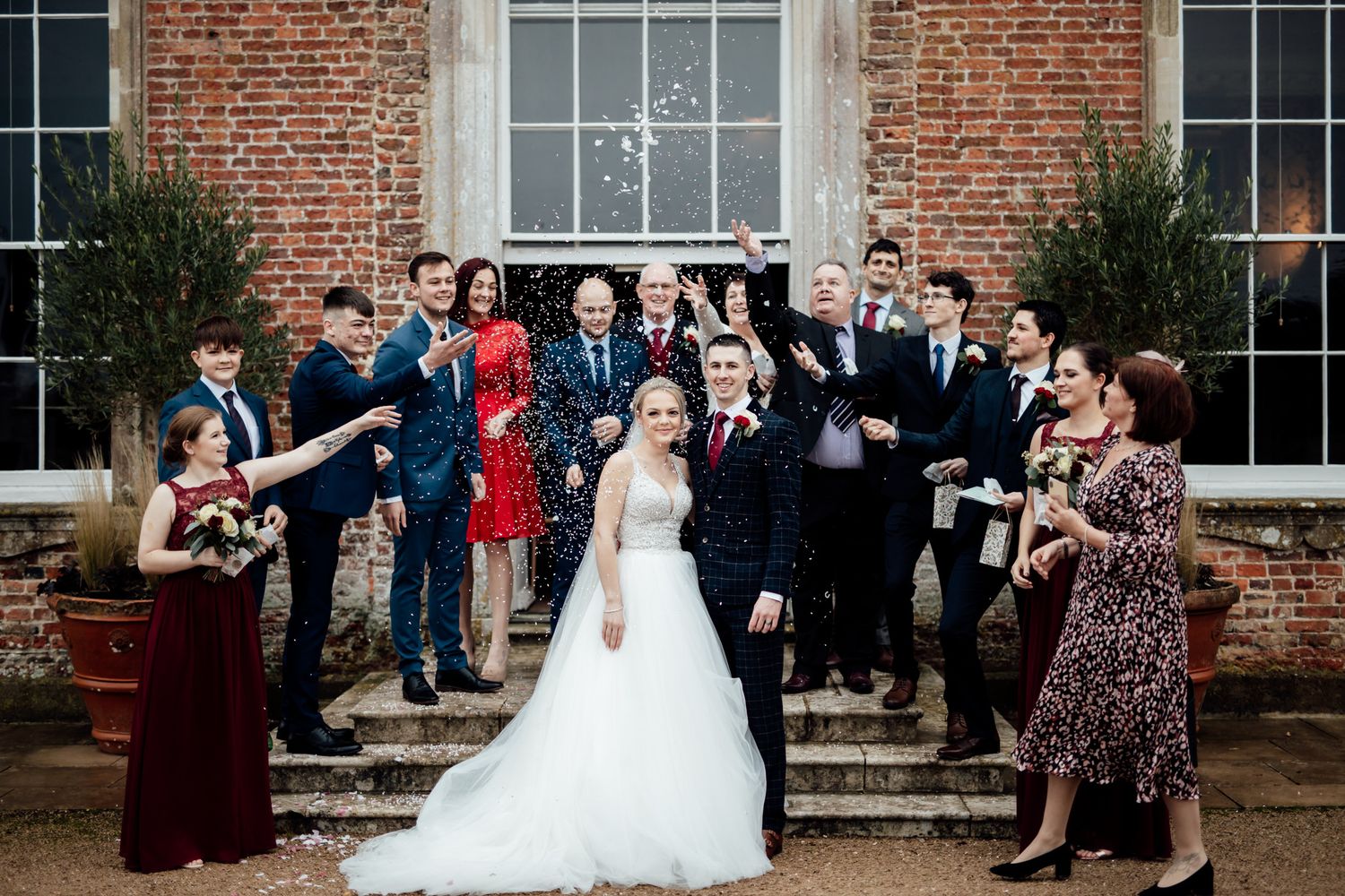 Wedding party and guests gathered outside brick mansion for group photos.