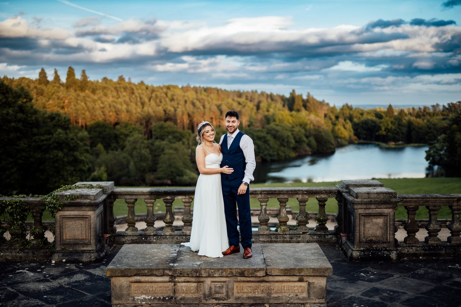 Wedding couple posing on stone balcony overlooking scenic lake and forest landscape.