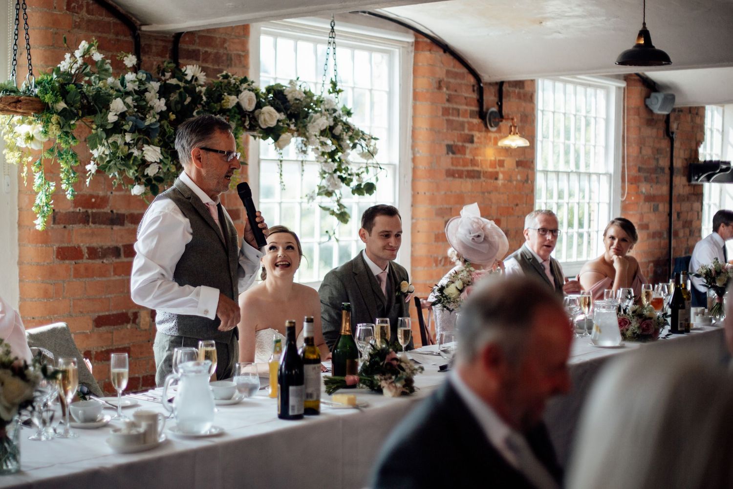 Long reception table setup in rustic brick venue with white linens and window lighting.