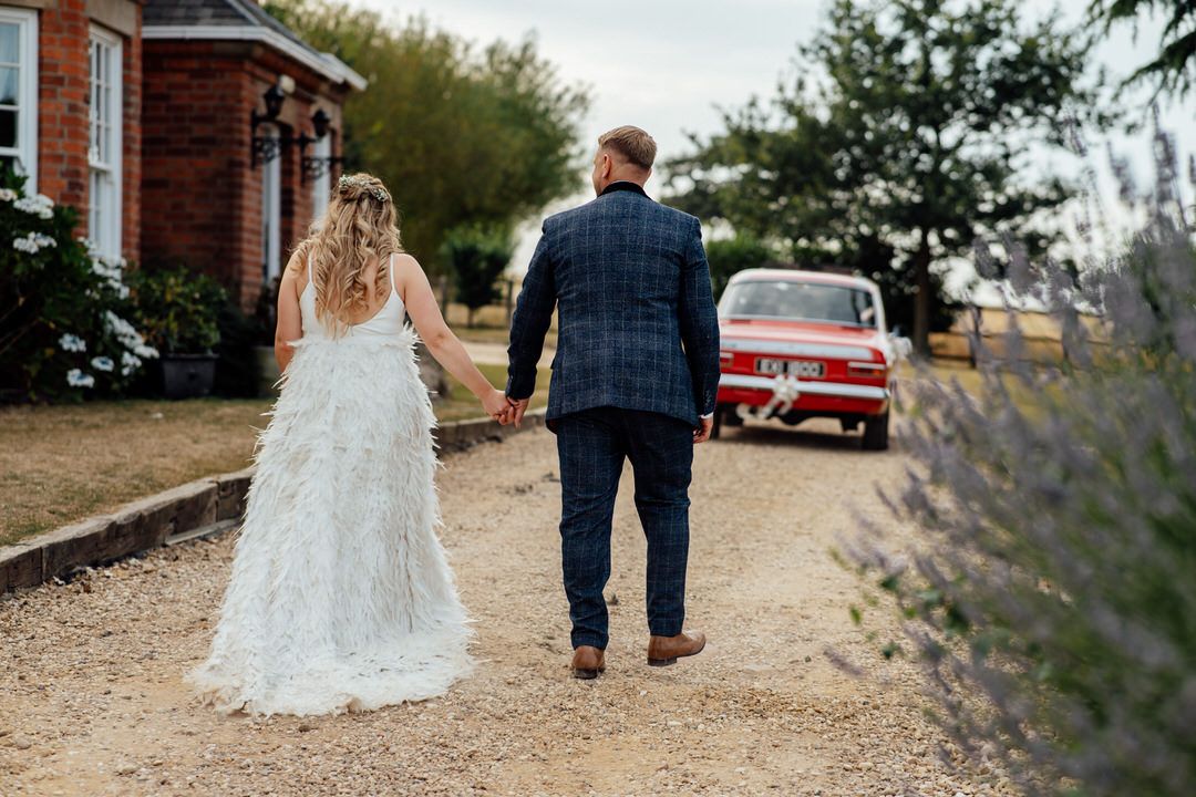 Wedding couple strolls along a rustic path lined with lavender and red brick buildings.