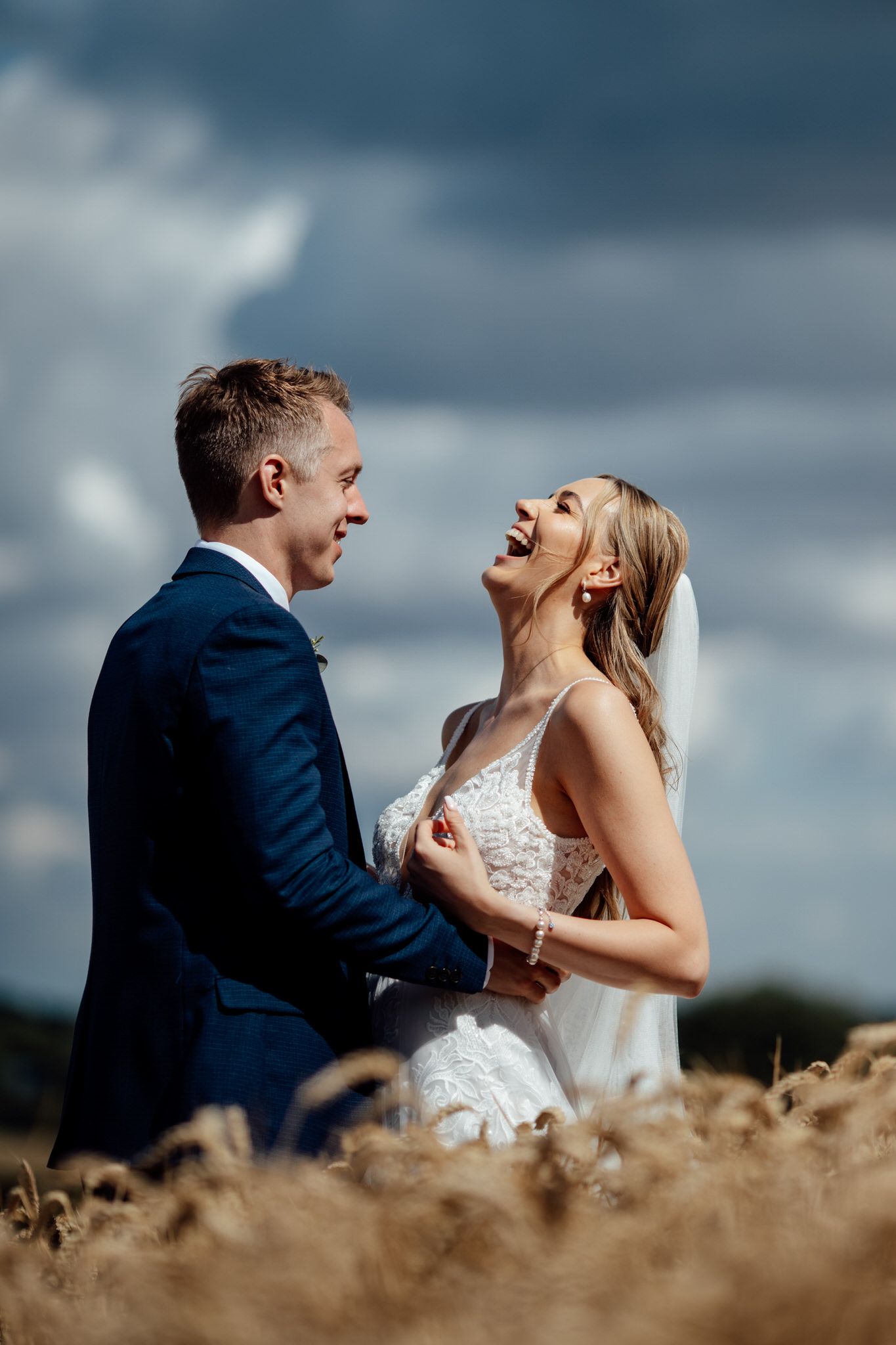 Joyful wedding couple laughing together in a golden wheat field against dramatic cloudy sky.